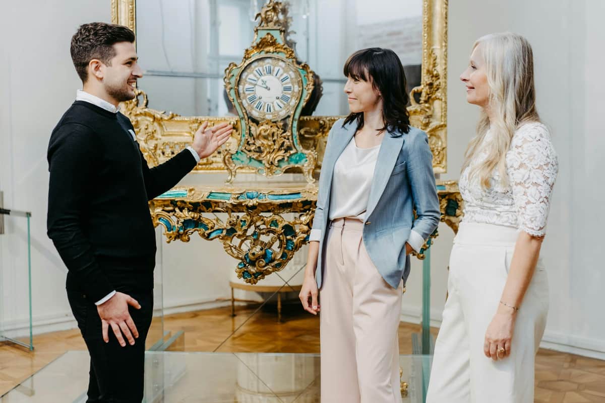 Two women stand near an ornate antique clock, with a third person gesturing towards them. The scene appears to be indoors.