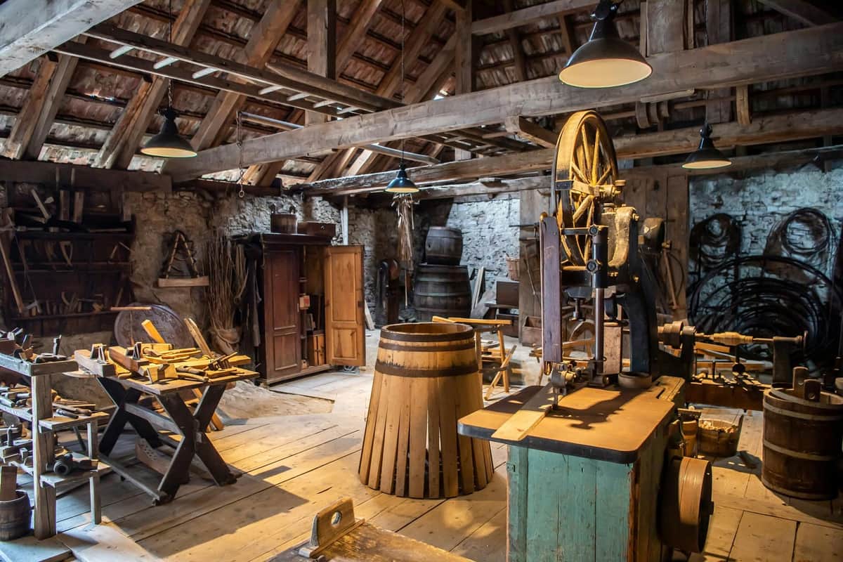 A rustic workshop filled with wooden tools, a barrel, workbenches, an old machine, and wooden beams under a high ceiling with lamps.