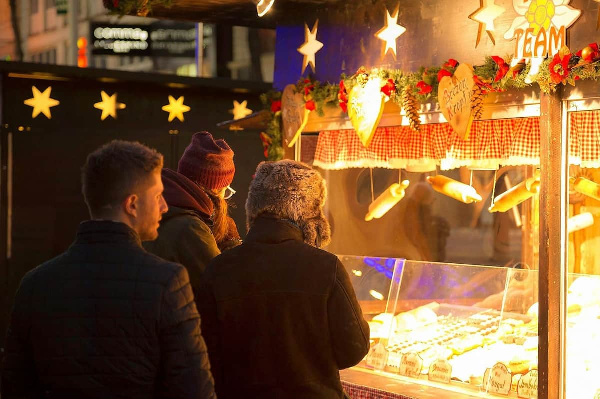 Viena: Visita guiada ao Mercado de Natal do Palácio de Schönbrunn + Degustação de strudel de maçã Três pessoas, vestidas com roupa de inverno, dão uma vista de olhos numa banca de um mercado ao ar livre com luzes decorativas e decoração com motivos festivos.