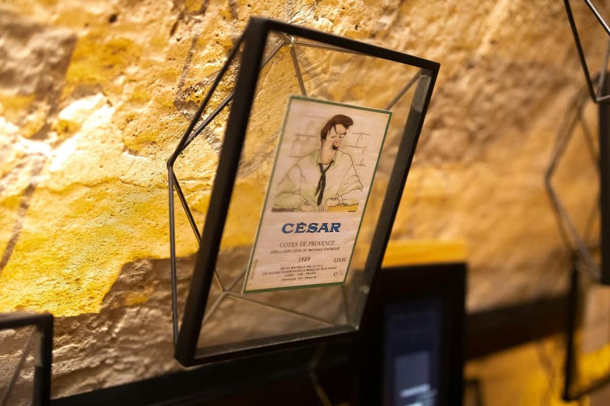 Les Caves du Louvre: French Wine Experience A framed illustration of a man with a bottle, labeled "César," displayed on a stone wall.