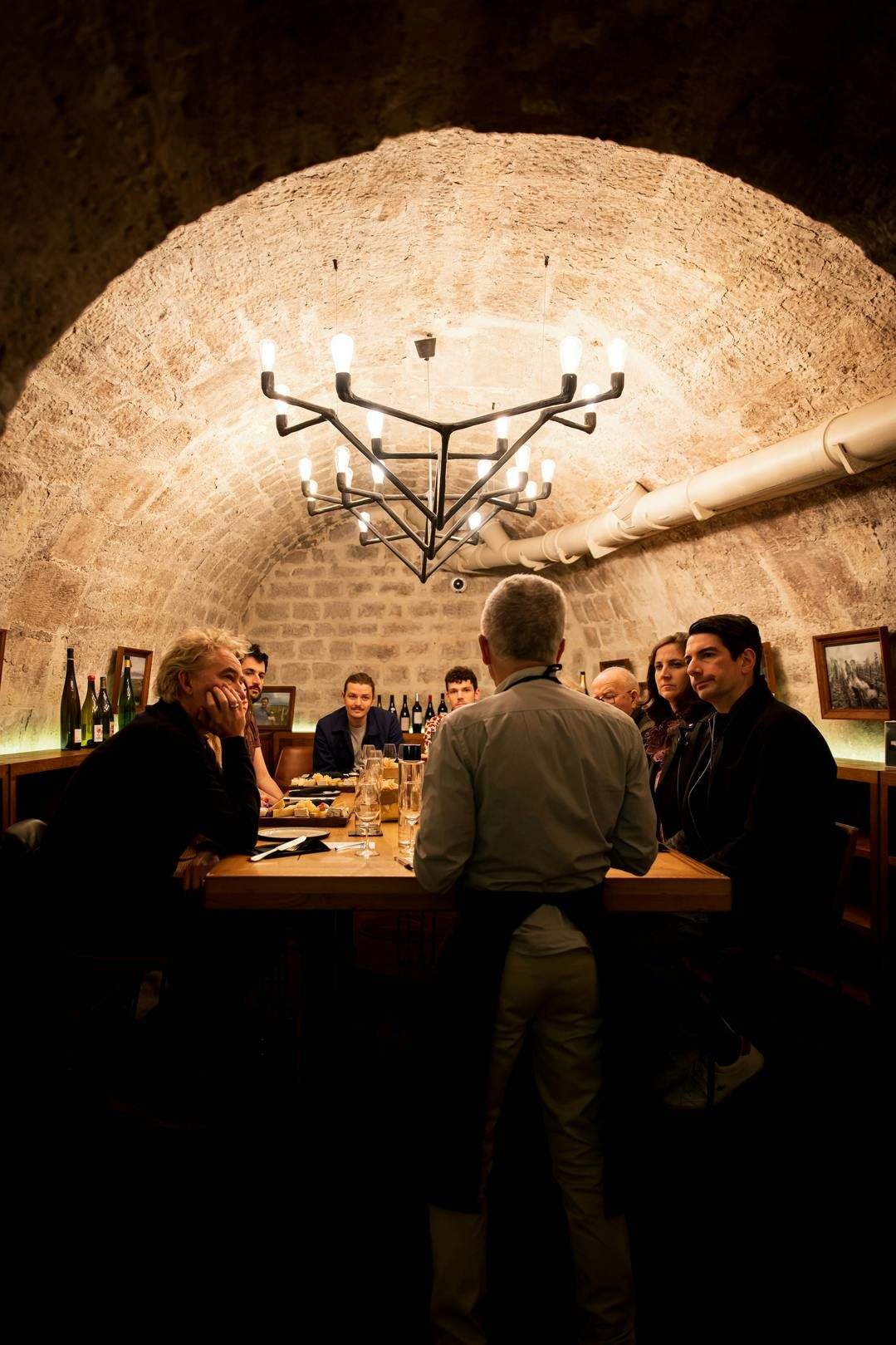 Les Caves du Louvre: Guided Wine and Cheese Tasting A group of people seated at a dining table in a stone-arched room, with a waiter standing at the table. A chandelier is above.
