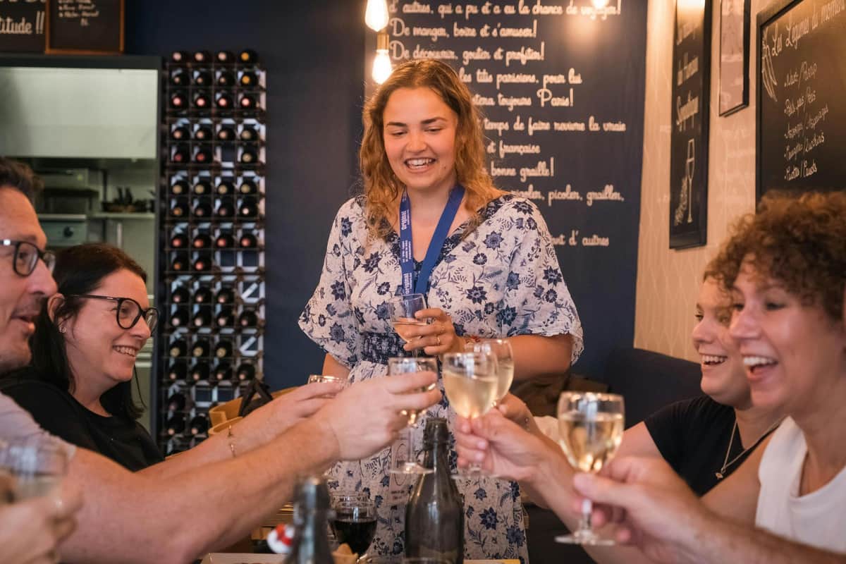 Montmartre Paris: French Bistrologie & Wine Tour A woman smiles while holding a glass of wine, surrounded by others toasting with wine glasses in a cozy setting with wine racks.