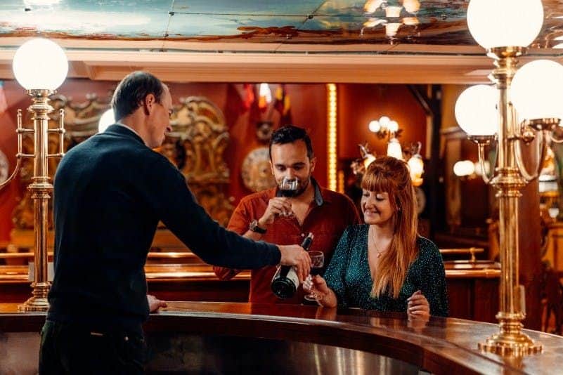 Le Hameau Duboeuf: Entry Ticket Three people at a bar: bartender pouring wine for a man and a woman. The background has warm lighting and ornate decor.