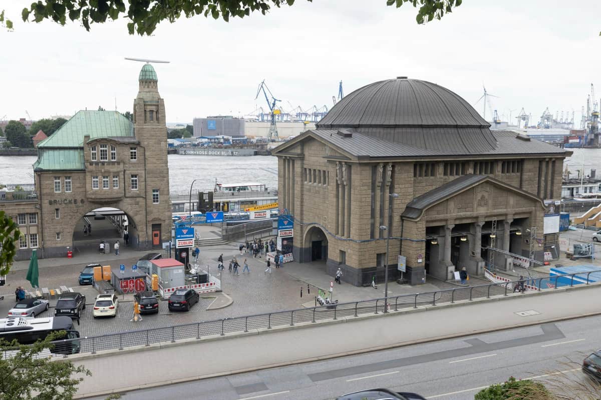 An urban scene featuring a large stone building by the waterfront, with cranes and industrial structures in the background. People and cars are visible.