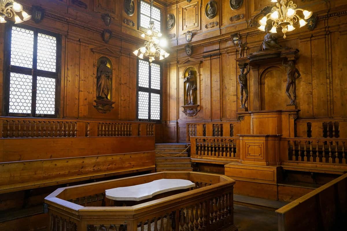 Wood-paneled room with statues on walls, chandeliers, a central marble-topped podium, and tiered wooden seating.