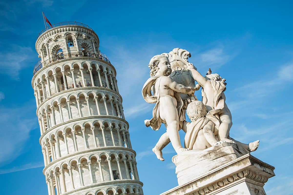 Pisa, San Gimignano & Siena: Day Trip from Florence + Siena City Tour + Lunch White marble statue of three cherubs in front of the Leaning Tower of Pisa under a clear blue sky.