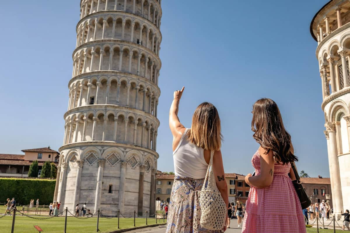 Pisa, Siena & San Gimignano: Day Trip from Florence + Lunch Two women stand near the Leaning Tower of Pisa, one pointing at it. A sunny day with a clear blue sky.
