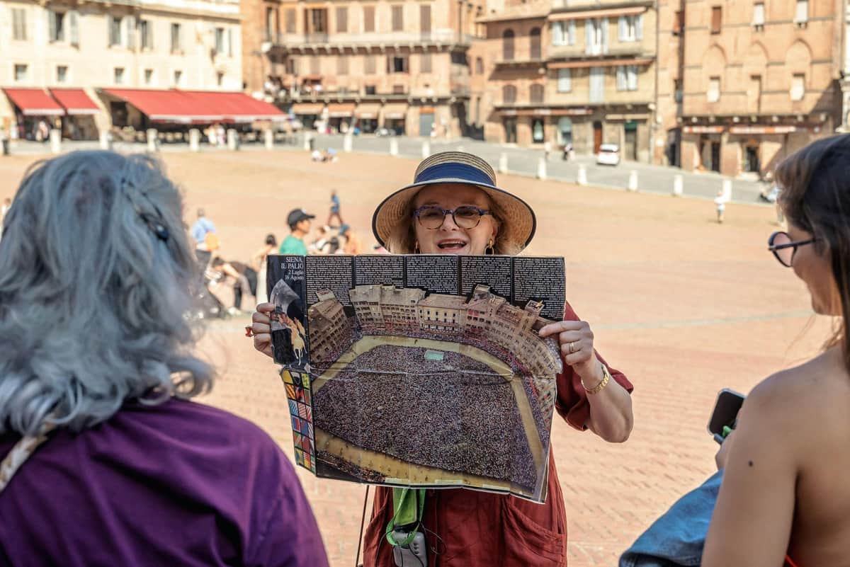 A person wearing a hat and glasses holds up a poster of a crowded square, surrounded by historic buildings, in an open plaza.