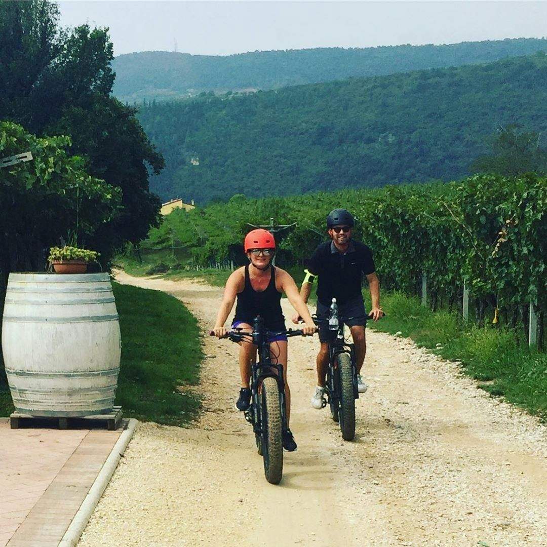 Valpolicella Vineyards: E-bike Tour with Wine Tasting Two people cycling on a gravel path through a vineyard, wearing helmets. Hills and greenery are visible in the background.
