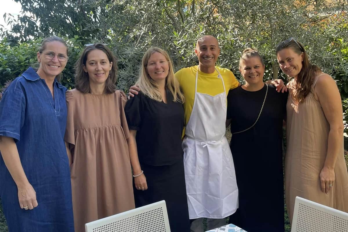 Emilia-Romagna: Pasta Making Class + Wine Tasting Group of five people standing outdoors, smiling. One person in a yellow shirt wears an apron. Trees and outdoor chairs are visible.