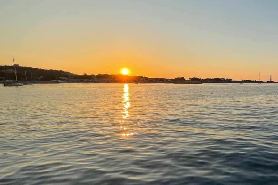 Palermo: Private Sunset Sailboat Tour + Dinner + Wine Sunset over a calm body of water with a reflection of the sun and a distant shoreline with trees and buildings.