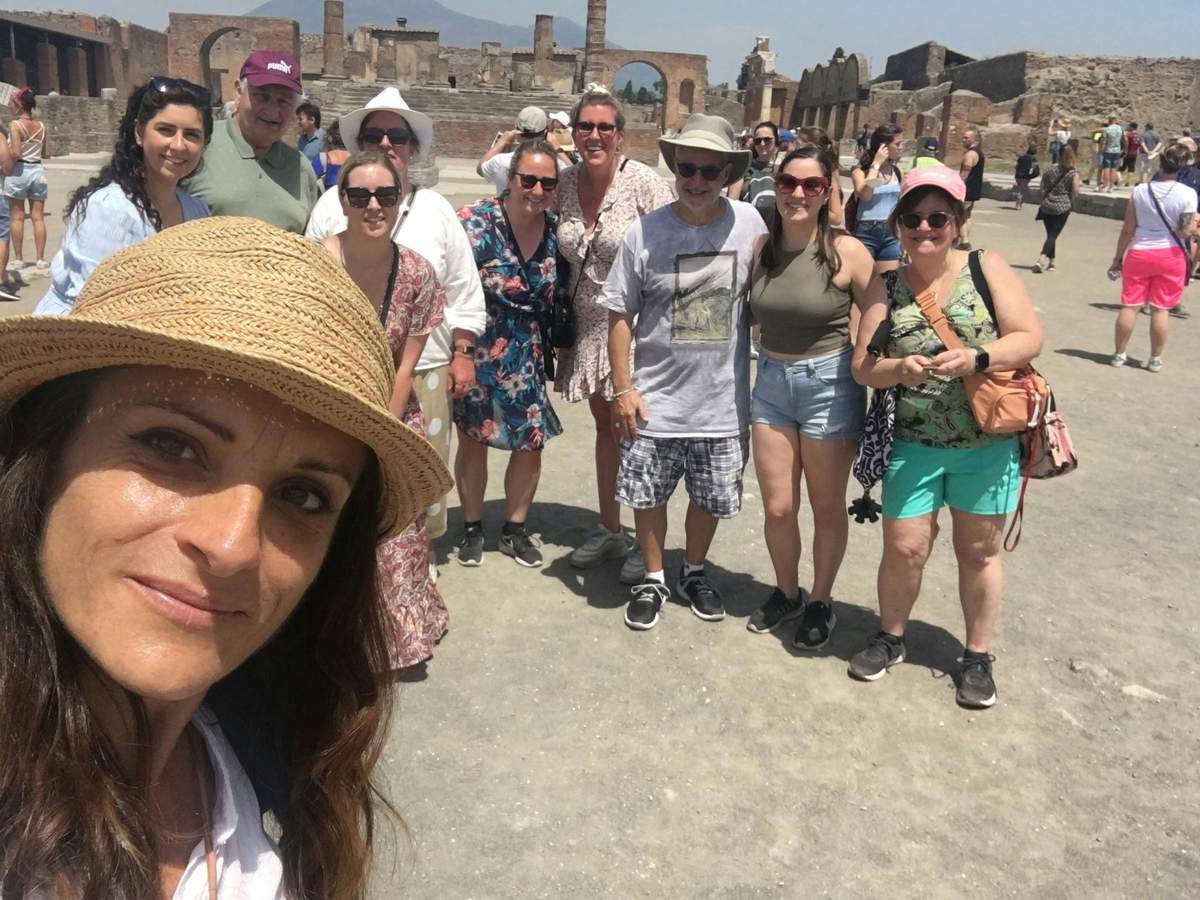 Pompeii: Skip The Line Ticket + High Speed Train from Rome Group of people smiling for a photo at an ancient ruins site on a sunny day. A person in a hat takes the selfie.