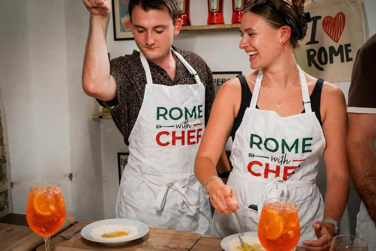 Two individuals in aprons, labeled "Rome with Chef," preparing dishes at a counter with drinks in the foreground.