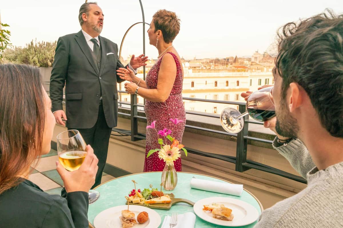 Rome: Trastevere Rooftop Opera + Aperitivo + Wine Three people at a rooftop setting with food and drinks on a table; two are conversing while one observes, holding a glass.