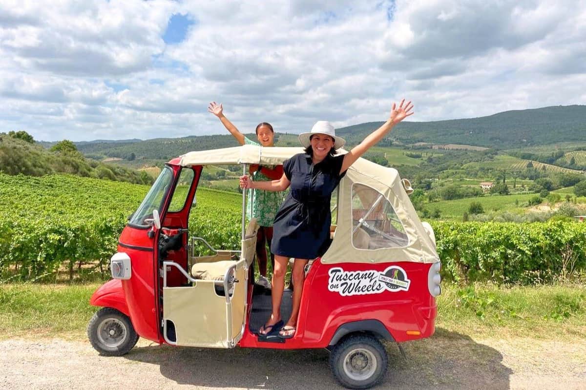 Tuscany: Private Guided Tuk Tuk Tour + Picnic + Olive Oil & Wine Tasting Two people cheerfully pose next to a red tuk-tuk in a vineyard landscape under a partly cloudy sky.