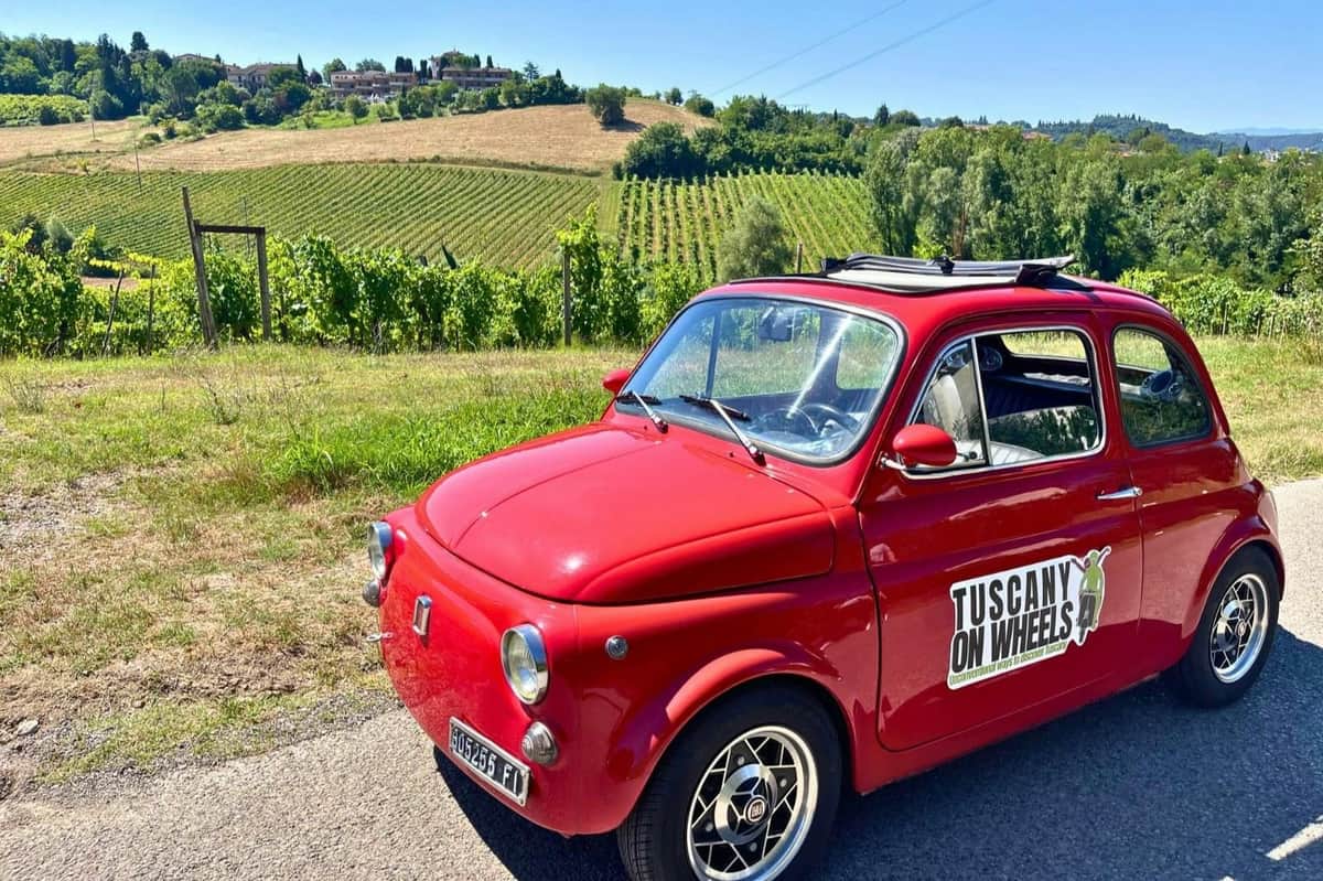 A red vintage car parked on a scenic road with vineyards and a hillside village in the background.