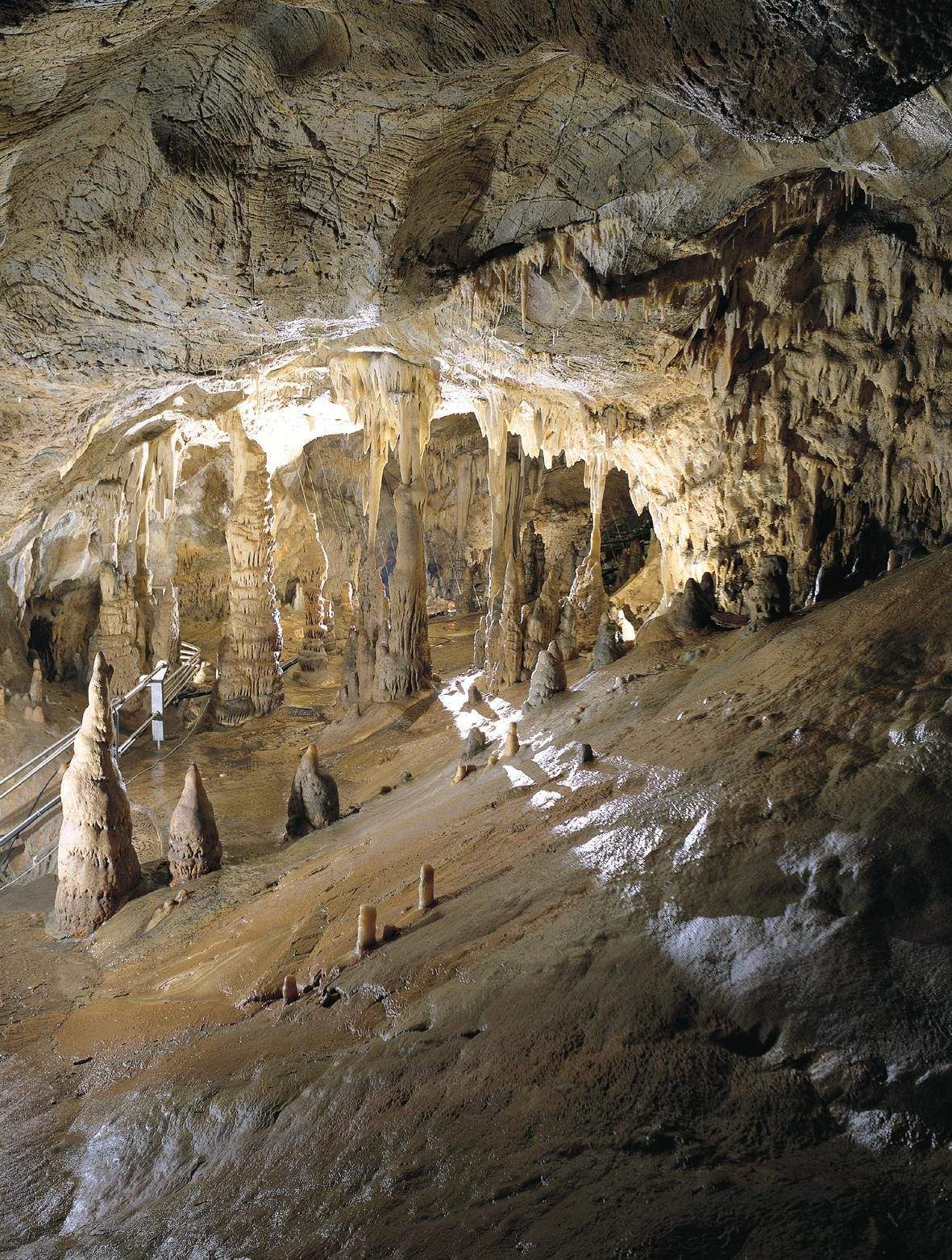 A cave with numerous stalactites hanging from the ceiling and stalagmites rising from the ground, with a walkway in the background.