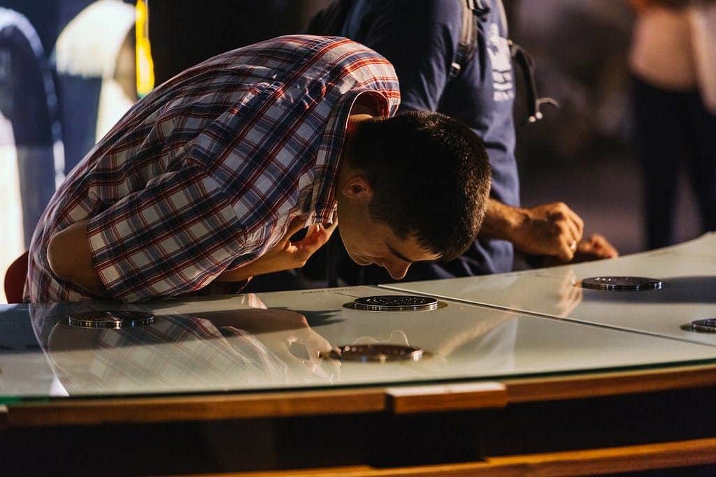 Cálem Caves: Guided Tour in Interactive Museum & Wine Cellars + Wine Tasting A man in a checkered shirt leans over a glass display, closely inspecting its contents. Another person stands nearby.