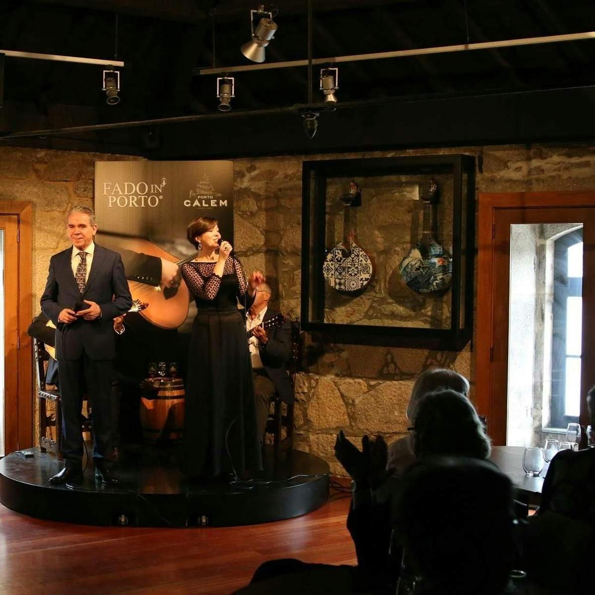 A woman sings on stage next to a man in a suit, with an audience watching in a rustic room adorned with ceramic vases.