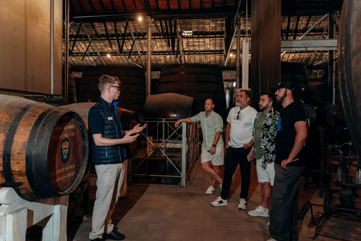 Porto: 2-Hr Private Tuk-Tuk Tour to Monastery and Cellars A group of five men in casual attire stand in a distillery or warehouse, surrounded by large wooden barrels, engaged in conversation.