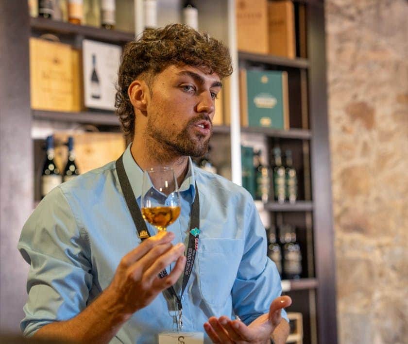 Porto City Tour Half-Day: Guided Visit with Wine Cellar and Tastings A person with a blue shirt and lanyard holds a glass of wine, speaking in a room lined with wine bottles and boxes.