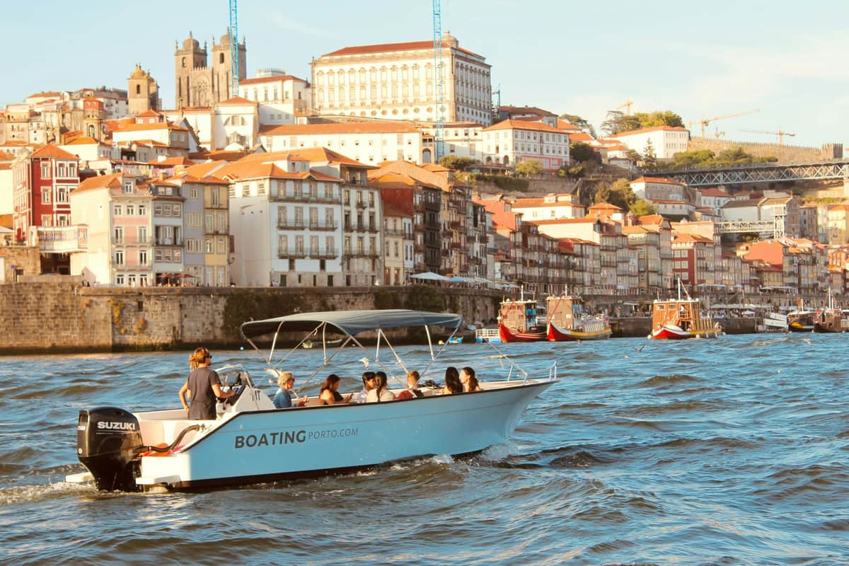 A boat with passengers on a river in front of a city with historic buildings and a cathedral on a sunny day.