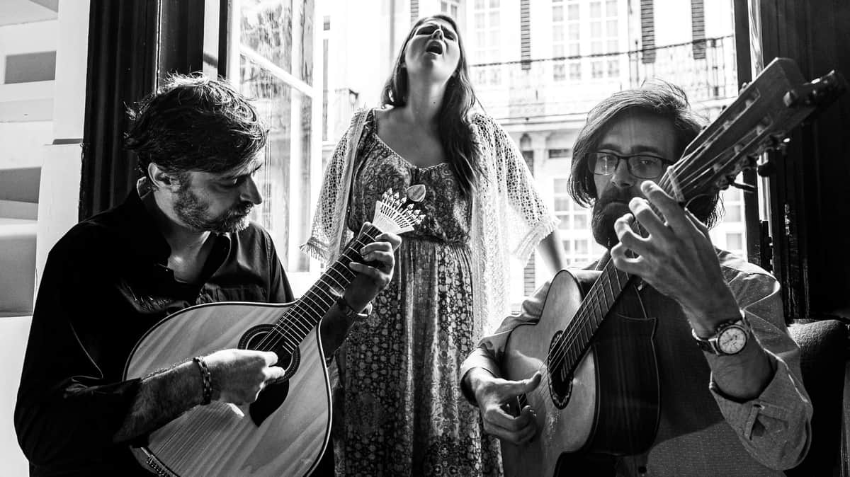 A man plays a guitar while a woman stands behind him singing passionately, in front of a window with shutters.