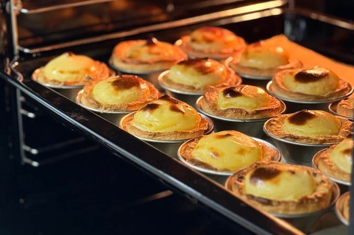 Rows of golden egg tarts baking in an oven, displayed on metal trays.