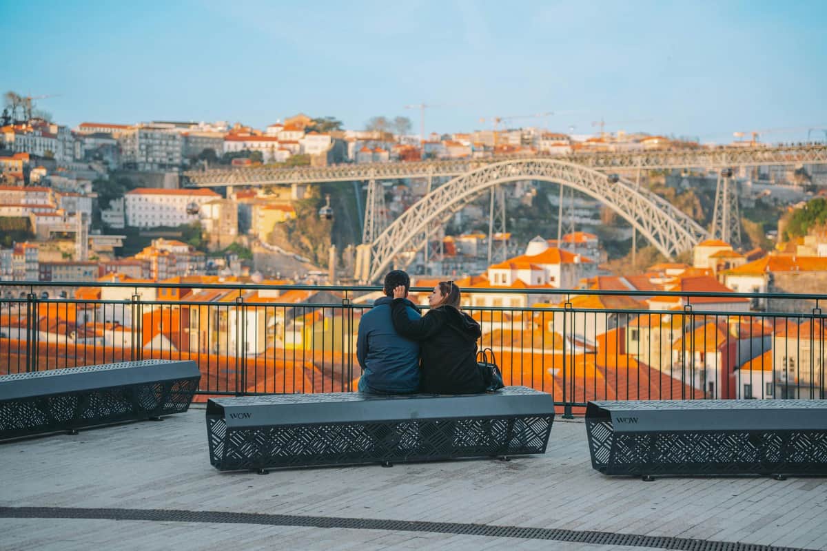 Two people sitting on a bench overlooking a cityscape with a bridge and red-roofed buildings under a blue sky.