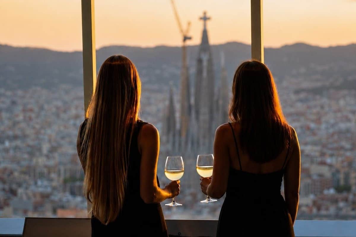 Mirador Torre Glòries: Guided Wine Tasting + Audio Guide Two women in black dresses, holding glasses of white wine, look out of a window at a cityscape featuring a tall church spire.