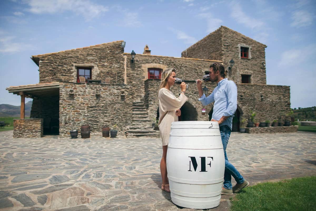 Martín Faixó | Sa Perafita: Guided Winery Tour A man and woman drink wine by a white barrel in front of a rustic stone building with stairs.