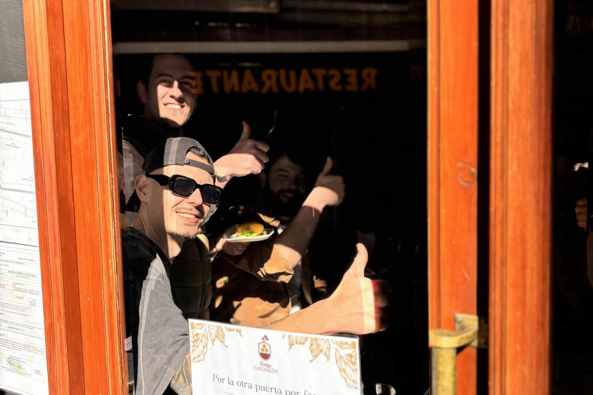 Three people behind a restaurant window giving thumbs-up, with one holding a plate. Menu and a sign visible on the window.