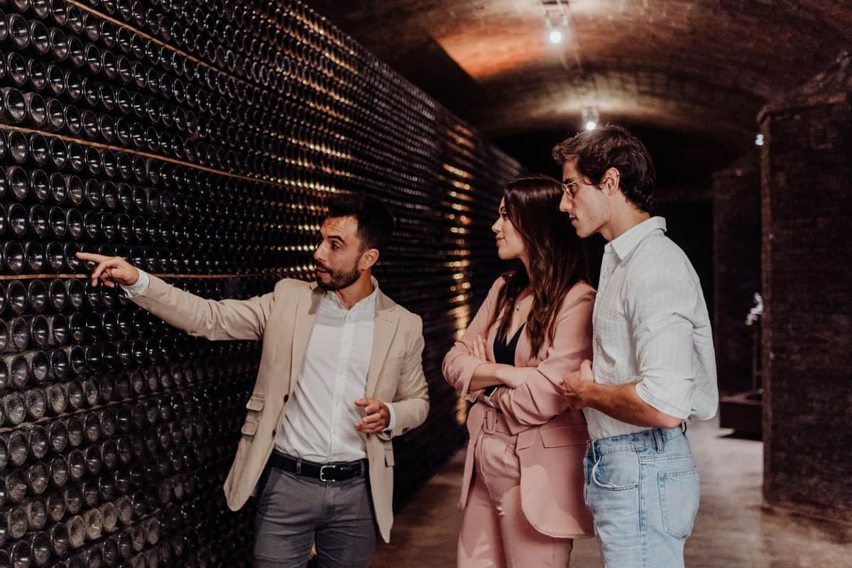 A man in a beige blazer explains something to a woman in a pink suit and a man in a white shirt in a wine cellar filled with bottles.