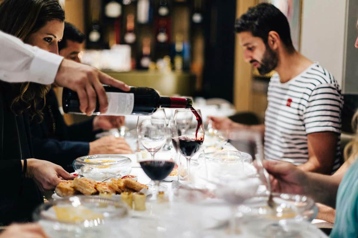 A person pours red wine into glasses at a dining table set with food, with people seated and dining in a softly lit room.