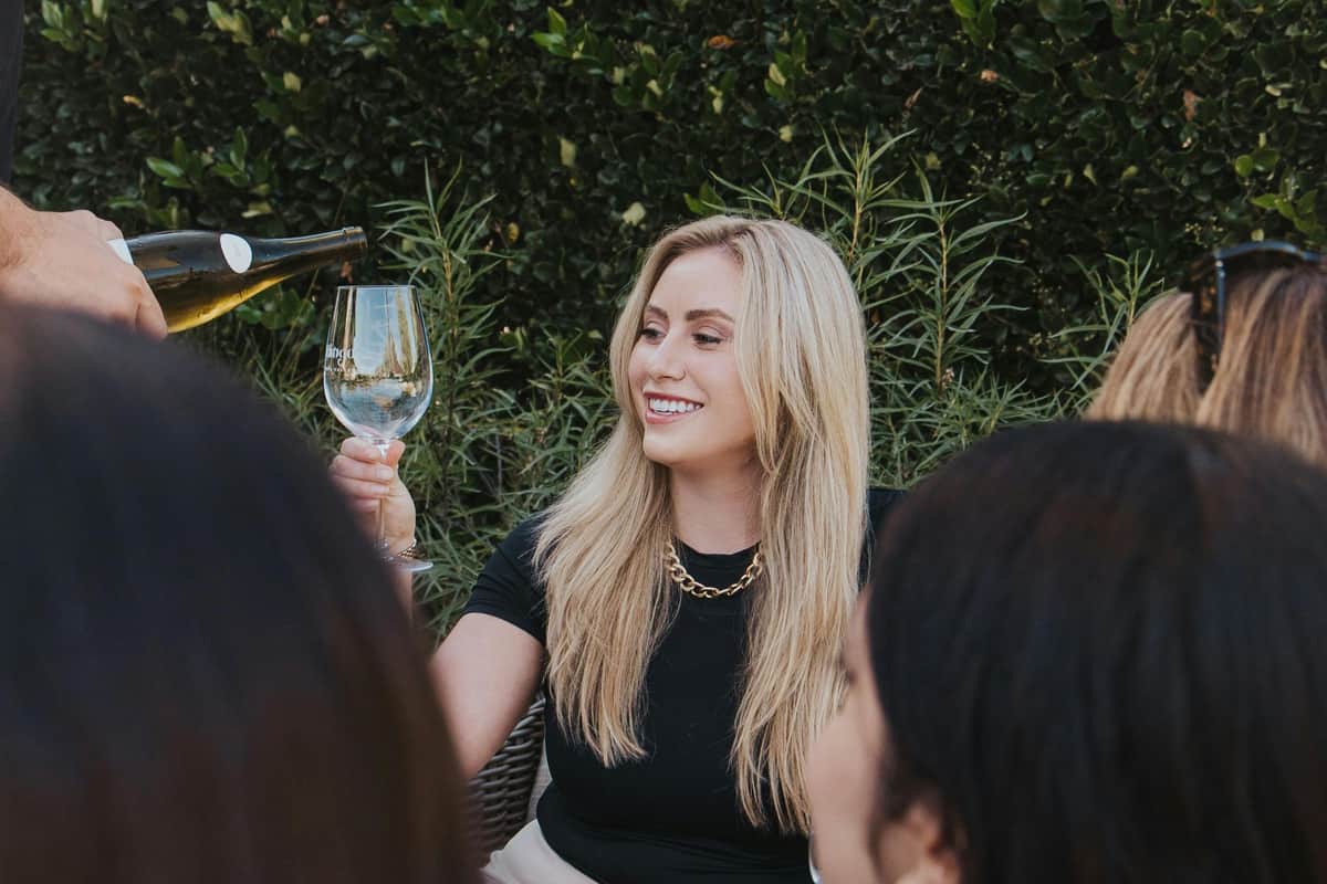 Napa & Sonoma: Guided Wine Tour from San Francisco A woman with long blonde hair smiles and holds up a wine glass as someone pours wine into it. Green foliage is in the background.