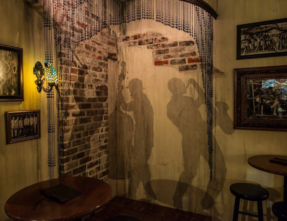 American Prohibition Museum: Entry Ticket or Guided Tour A dimly lit room with shadow murals of jazz musicians on a brick wall, adorned with beaded curtains and vintage decor.