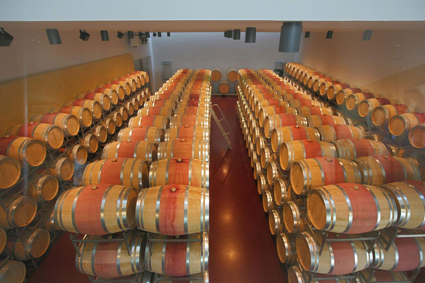Rows of stacked wooden wine barrels in a large, modern cellar with overhead lighting and a central aisle with a ladder.