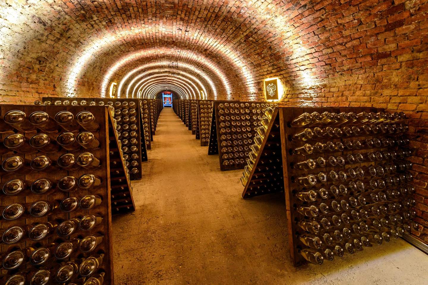 Schlumberger Kellerwelten A brick tunnel filled with racks of wine bottles, lit by warm lights, creating a vanishing point effect towards a distant door.