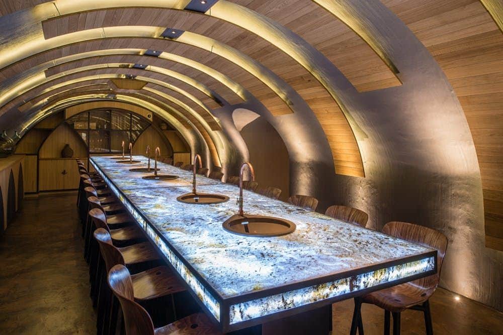 A modern bar with an arched wooden ceiling, marble countertop, and wooden chairs, illuminated by warm, integrated lighting.