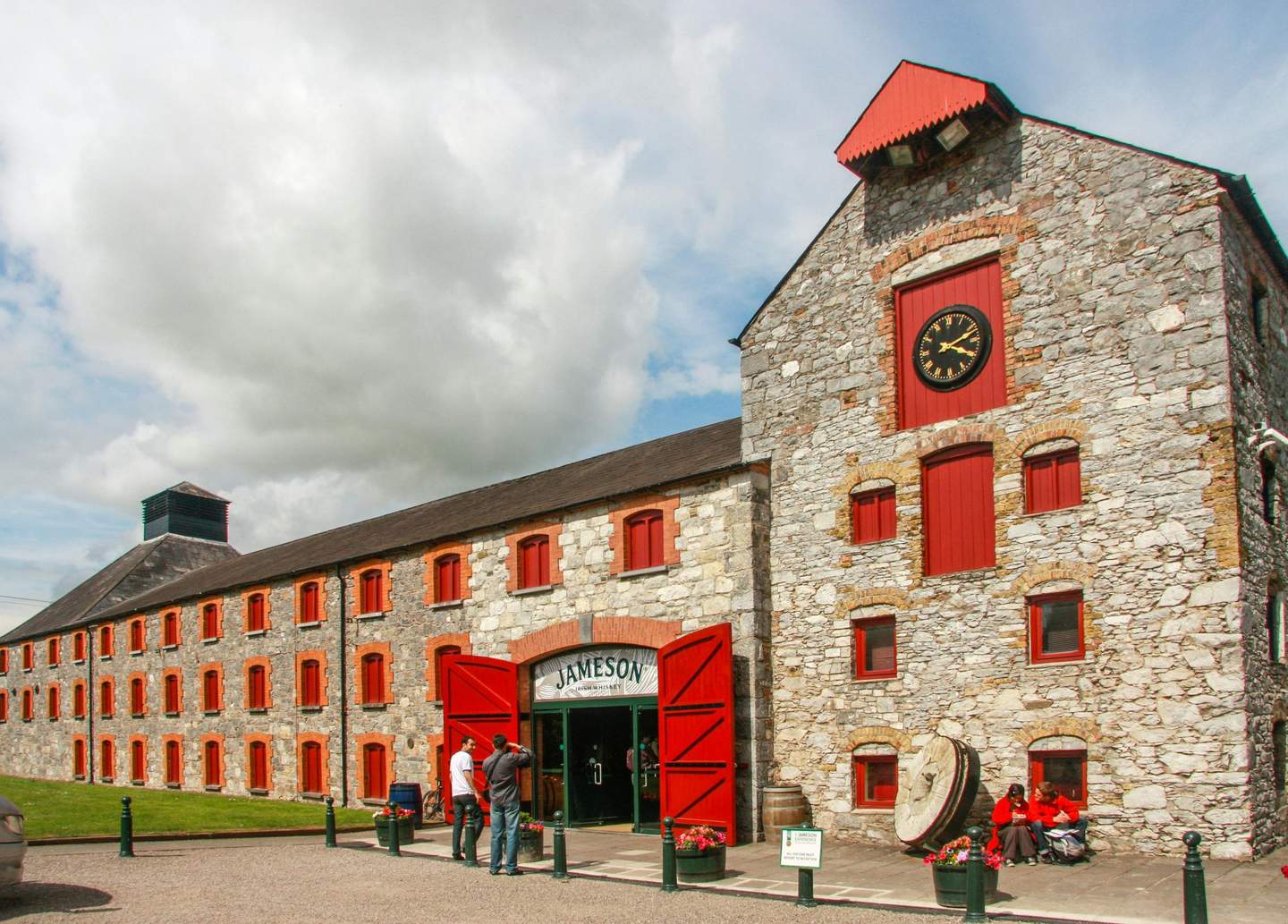 Stone building with red doors and windows, labeled "Jameson." Clock above the entrance. Three people sit near the entrance.