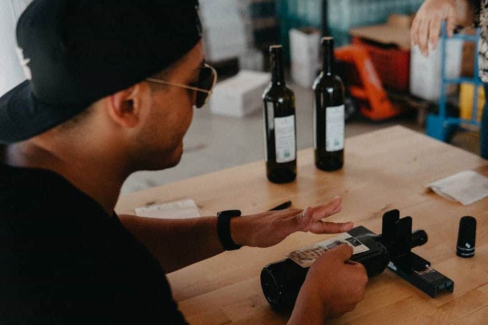 Person in a cap and sunglasses holding a bottle near a label machine, with two wine bottles on a wooden table.