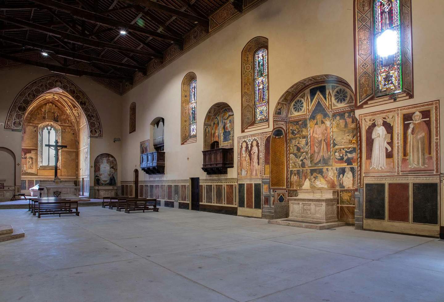 Interior of a historic church with wooden ceiling, stained glass windows, frescoes, pews, a large cross, and a stone altar.
