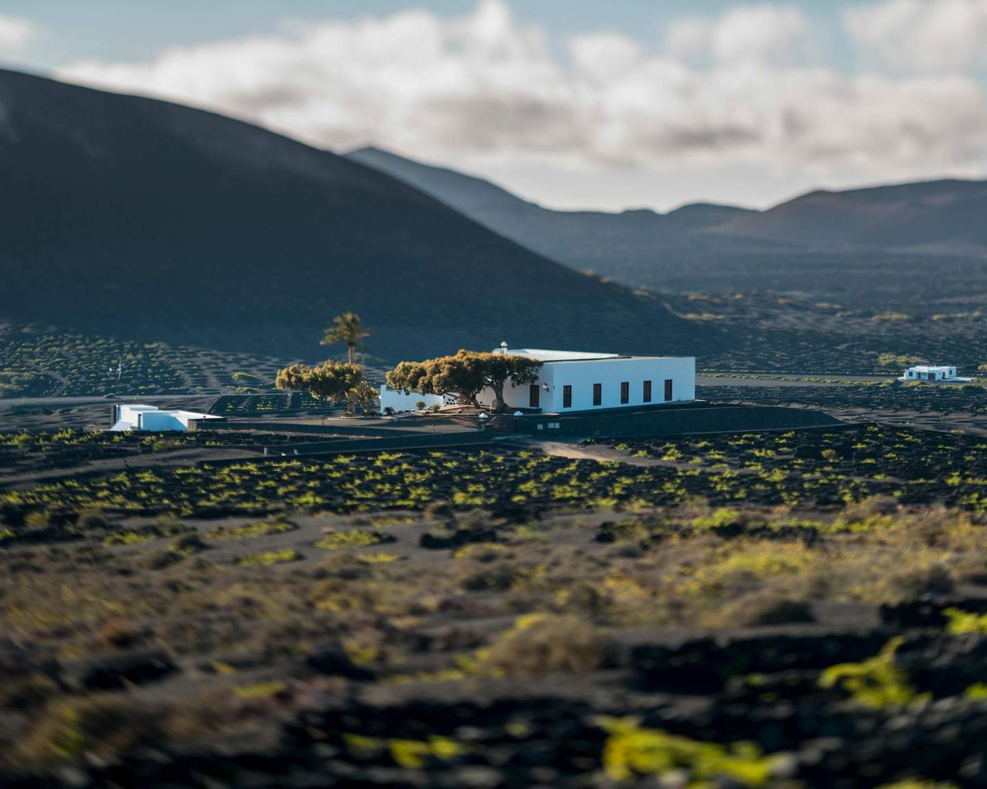 White buildings surrounded by sparse vegetation in a vast, rocky landscape with hills in the background under a cloudy sky.