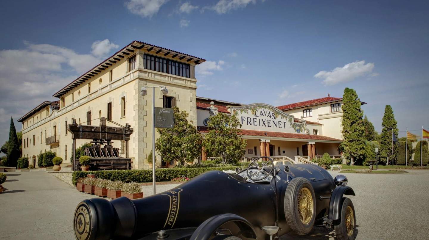 Exterior of Cavas Freixenet building with a vintage car parked in front, greenery, and clear blue sky.