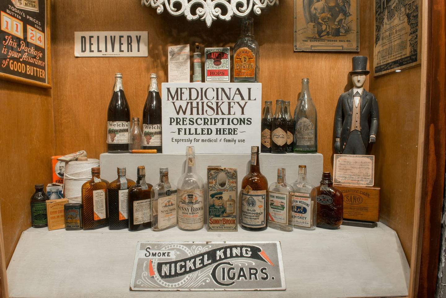 Display case with vintage liquor bottles, a "Medicinal Whiskey Prescriptions Filled Here" sign, and a wooden figurine of a man in a top hat.
