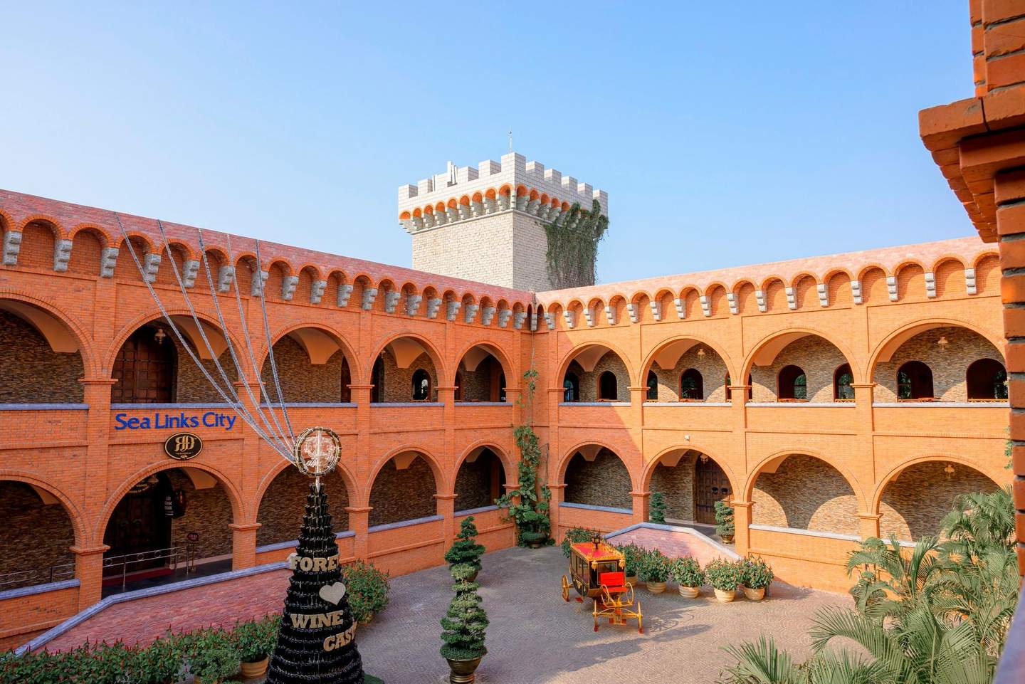 RD Wine Castle A courtyard with red brick architecture, a decorative wine display, a vintage carriage, and plants under a blue sky.