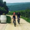 Two people cycling on a gravel path through a vineyard, wearing helmets. Hills and greenery are visible in the background.